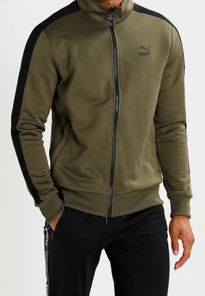 Mężczyzna w oliwkowej kurtce Puma na zamek z czarnymi paskami na rękawach oraz czarnych spodniach z bocznym logo, ręce swobodnie wzdłuż ciała.