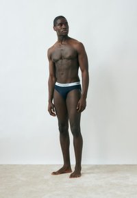 Tume naha tooniga meesmodell seisab paljajalu, seljas tumesinised aluspüksid valge vöökohtaga, millel kiri "Emporio Armani", taustal tavaline hele taust.