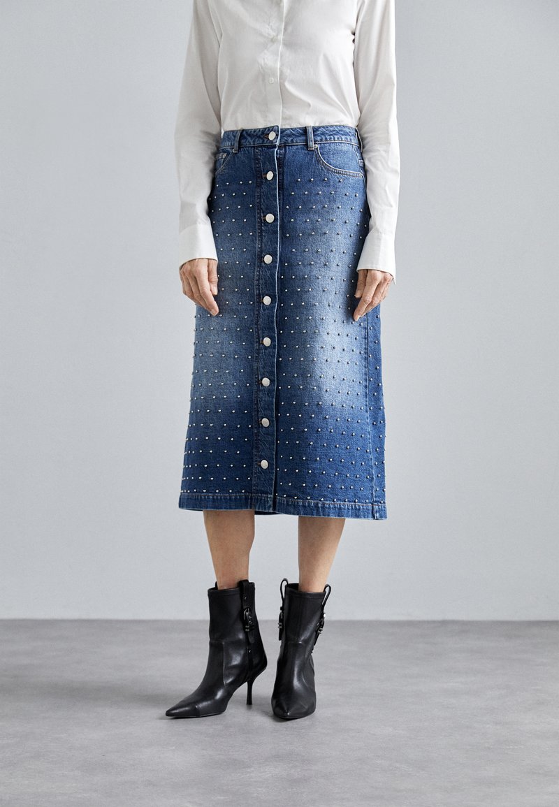 Jeansrock mit taillierter Passform, knielang, verziert mit silbernen Nieten und vorne mit Knopfverschluss; kombiniert mit schwarzen Knöchelstiefeln.