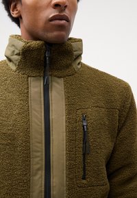 Fleecejacke in Olivgrün mit strukturiertem Finish, hohem Kragen, Reißverschluss und einer Brusttasche mit schwarzem Hardware-Detail.
