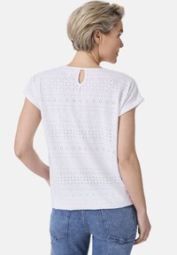 Weißes Kurzarmoberteil mit strukturierten Lochmustern, einem Rückenschlitz und einem lässigen Schnitt, kombiniert mit blauen Jeans.