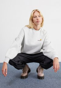 Jasnoszary sweatshirt z okrągłym dekoltem i haftowanym logo, w zestawieniu z luźnymi czarnymi spodniami cargo oraz beżowymi butami na sznurówki z grubą podeszwą.