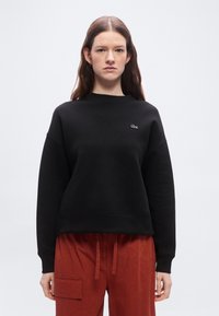 Γυναίκα με μα κριά καστανά μαλλιά φοράει μαύρο φούτερ με λογότυπο στο στήθος και φαρδιά κόκκινα παντελόνια με φόντο απλό λευκό.