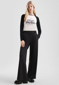 Blonde Frau trägt ein schwarz-weißes Langarmhemd mit dem Text "High Street", schwarze schmal gestreifte weite Hosen und schwarze klobige Schuhe.