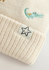 Crème kleurige gebreide beanie met een omgeslagen rand, voorzien van een klein geborduurd groen krokodil en een genaaide tag met een lachende ster.