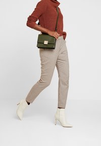 Pull en maille marron à col montant, sac bandoulière en cuir vert avec un fermoir carré doré, pantalon à carreaux beige, et bottines blanches à bout pointu.