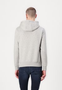 Un hombre de pie, de espaldas a la cámara, lleva puesto un sudadera con capucha gris claro y jeans oscuros frente a un fondo liso claro.