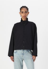 Giacca nera corta con colletto alto, dotata di zip frontale, vestibilità ampia e maniche lunghe, abbinata a jeans azzurri chiari.