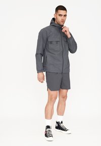 Graue wasserfeste Jacke mit zwei Brusttaschen, kombiniert mit passenden Shorts. Das Modell trägt schwarze hohe Sneakers mit weißen Akzenten.