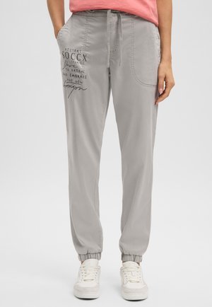 Frau trägt hellgraue Jogginghosen mit elastischen Bündchen und schwarzem Schriftzug am linken Oberschenkel, kombiniert mit weißen Turnschuhen und einem rosa Oberteil.