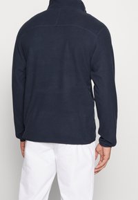 Mörkblå fleece-pullover med hög krage, långa ärmar och flatlocksömmar. Har en slät yta och fållade muddar.