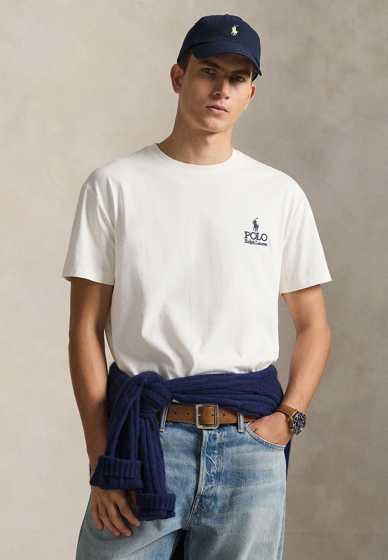 Λευκό βαμβακερό t-shirt με μικρό ναυτικό λογότυπο, σετ με ανοιχτό γαλάζιο τζιν και ναυτικό πουλόβερ δεμένο στη μέση. Καφέ ζώνη και ρολόι.