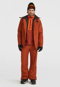 Outdoor ski outfit in orange and brown, featuring a zippered jacket, insulated pants, and gloves, paired with a black beanie and sunglasses.