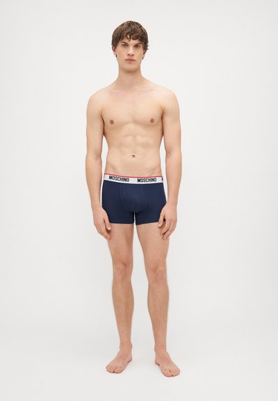 Boxers bleu marine avec une ceinture élastique arborant un logo rouge et noir. Le tissu semble lisse et ajusté. Pieds nus visibles.