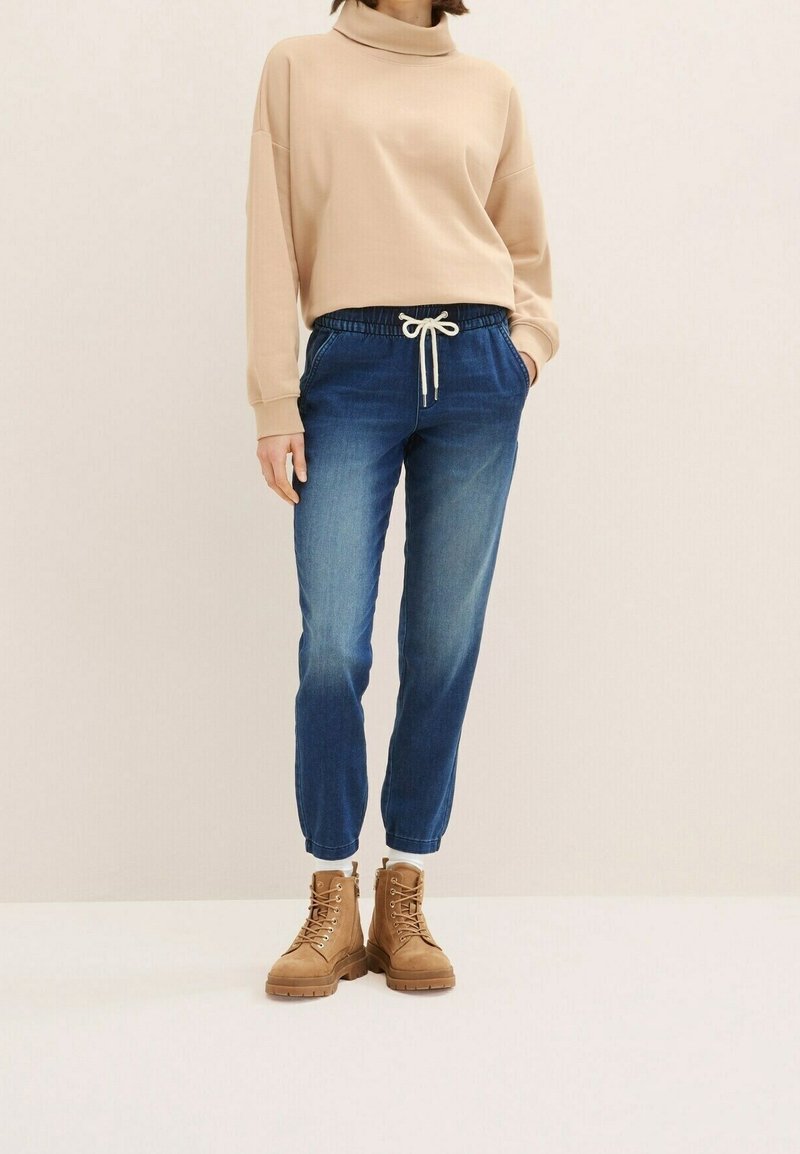 Personne portant un sweat-shirt à col roulé beige, un jean bleu à cordon de serrage et des bottines montantes beige à lacets sur un fond uni.