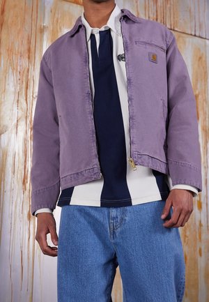 Homme portant une veste zippée violette par-dessus un maillot de rugby rayé bleu marine et blanc et un jean bleu, debout devant un mur texturé en bois.