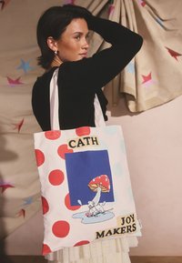 Lühikeste tumedate juustega naine, kes kannab musta tuba, hoiab valget käekotti punaste täppide ja seene illustratsiooniga, tekstiga „CATH JOY MAKERS".