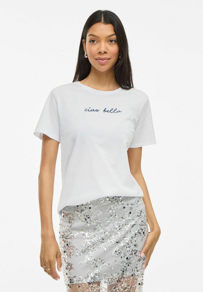 T-shirt en coton blanc à manches courtes et encolure ronde, avec "ciao bella" en écriture cursive marine. Porté avec une jupe en sequins argentés.