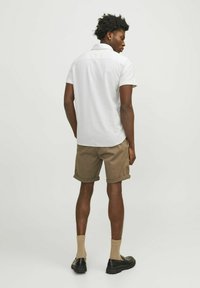 Camisa blanca de manga corta con una textura sutil, combinada con pantalones cortos marrones enrollados. Footwear negro y calcetines color beige completan el conjunto.
