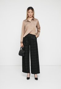 Camicia button-up in satin color marrone chiaro, pantaloni a gamba larga neri e bianchi a trama, borsa nera con superficie patterned, tacchi neri lucidi.