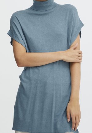 Persoon draagt een loszittende korte mouw blauwe coltrui-jurk met één arm over het lichaam gekruist.