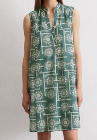 Ärmellös, knälång klänning i grönt med ett geometriskt vitt mönster. Har en kraglös halsringning och en avslappnad passform. Mjukt tyg.