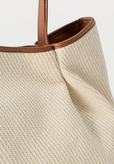 Κοντινή λήψη μπεζ υφασμένης τσάντας ώμου με καφέ δερμάτινα περιλαίμια και λαβές, που αναδεικνύει την υφή και τις λεπτομέρειες της ραφής.