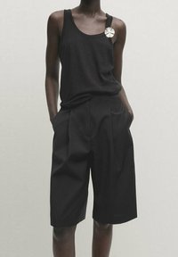 Personne portant un haut sans manches noir avec une broche métallique et un short noir ample coupé au genou, les mains dans les poches.