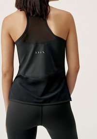 Svart atletisk linne med ett meshnät på axlarna, har en slät textur och ett centrerat vitt logotypdetalj på baksidan. Kombineras med svarta leggings.