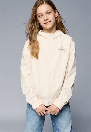 Giacca a zip color crema chiaro con cappuccio, polsini elastici e vita elasticizzata. Presenta un piccolo logo grigio sul lato sinistro del petto. Indossata con jeans blu.