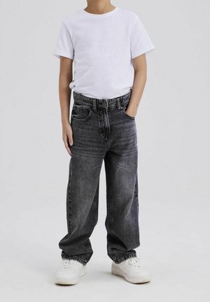 Dítě nosící jednoduché bílé tričko, volné černé vyprané džíny s rukama v kapsách a bílé tenisky na jednoduchém pozadí.