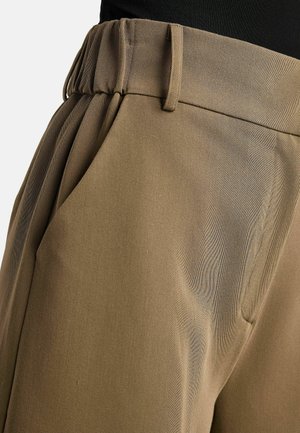 Primo piano di pantaloni beige a vita alta con vita arricciata, passanti per cintura, tasca laterale e tessuto dalla texture liscia.