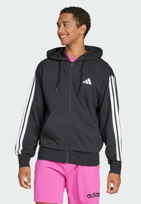 Hoodie preto com fecho, feito de tecido macio, apresentando riscas brancas nas mangas e um pequeno logótipo branco no peito. Capuz ajustável com cordão.