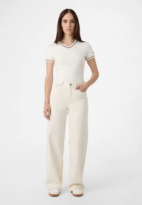 Dopasowany biały top z dekoltem w serek i czarną lamówką, połączony z kremowymi spodniami o wysokim stanie i szerokich nogawkach. Modelka ma na sobie jasne buty.