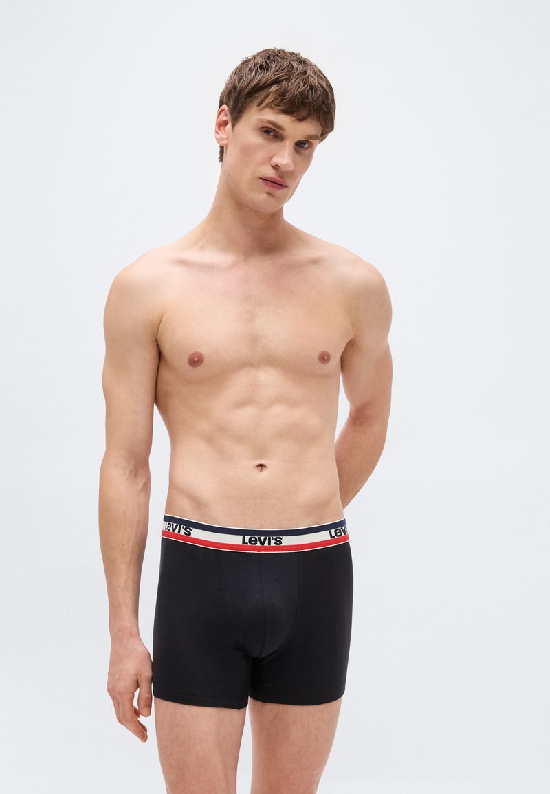 Shirtless man with short brown hair wearing black Levi's boxer briefs, standing against a plain light background with hands behind back.