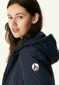 Marineblauwe geïsoleerde jas met een capuchon, voorzien van een rond logo-patch op de linkermouw. Zachte textuur en een gestroomlijnd ontwerp.