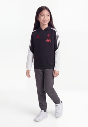 Dziewczynka stoi i się uśmiecha, ubrana w czarno-białą bluzę z kapturem Liverpool FC, ciemnoszare spodnie i białe trampki na białym tle.