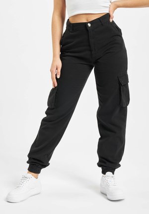 Femme portant un pantalon cargo noir avec des poignets élastiques et des baskets blanches, posant avec une main sur la hanche sur un fond blanc.