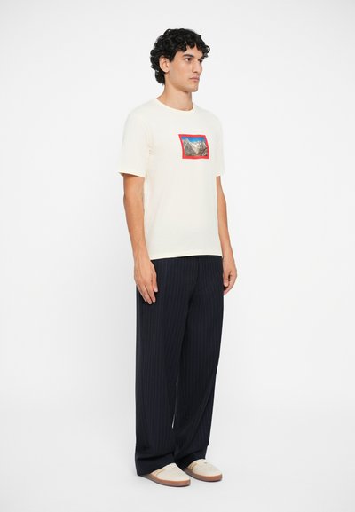 Cremefarvet kortærmet t-shirt med et rektangulært bjerggrafik i rød kant. Sammen med mørke stribede bukser og lyse sko.