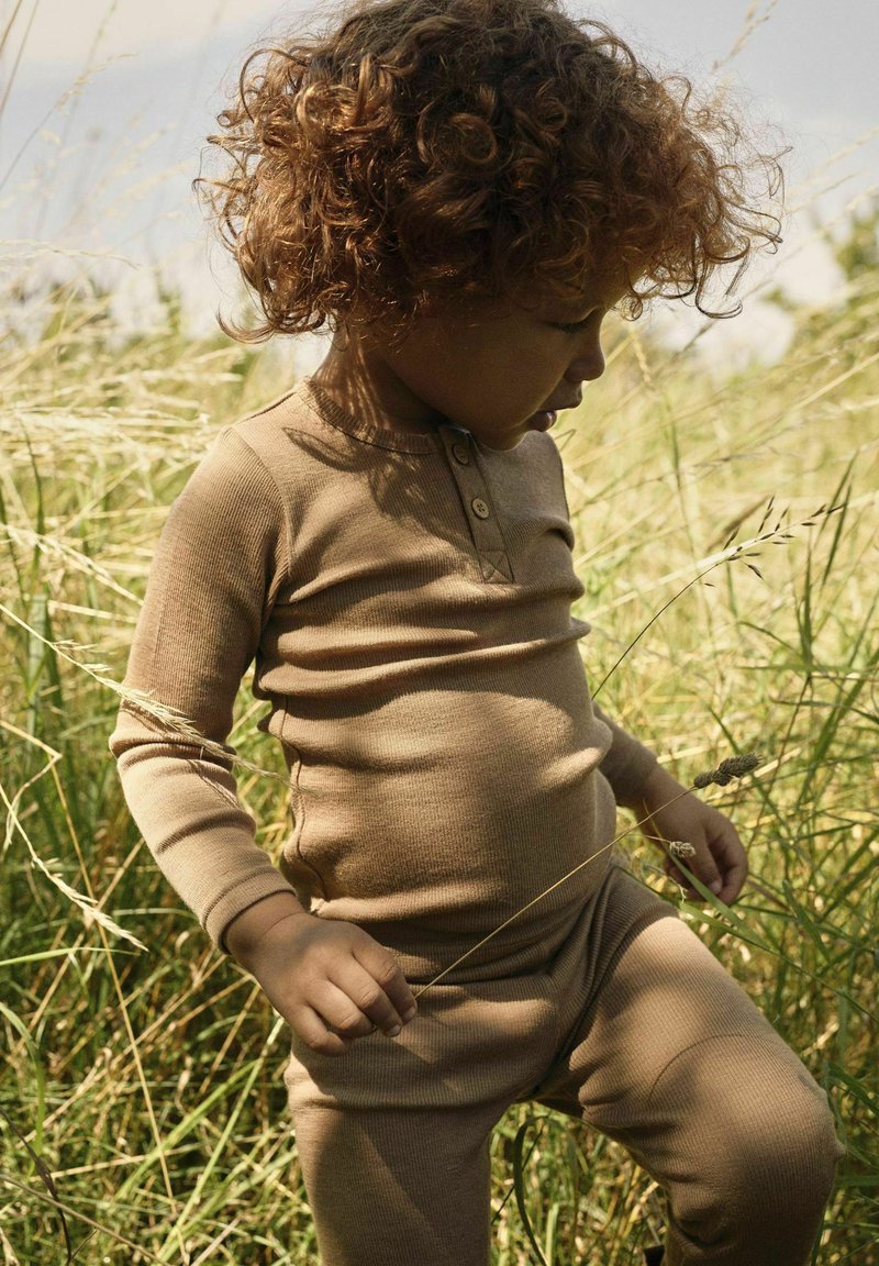 Jeune enfant aux cheveux bouclés portant une tenue beige, se tenant dans un champ herbeux, tenant des tiges d'herbe sèches, regardant vers le bas.