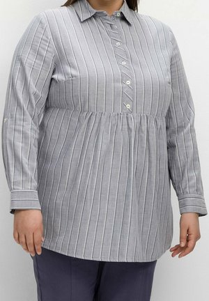 Kobieta nosząca szarą i białą pionowo prążkowaną bluzkę na guziki z długim rękawem i marszczonym paskiem w talii, zestawioną z ciemnymi spodniami.