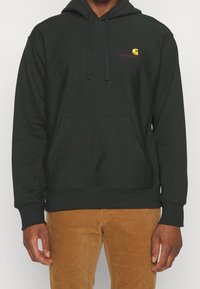 Felpa con cappuccio in tessuto verde scuro con tasca frontale. Presenta un piccolo logo rosso e giallo sul lato sinistro del petto. Polsini texturizzati.