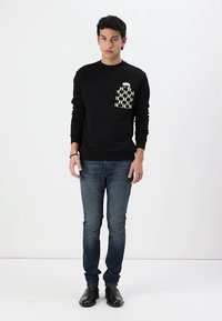 Sort sweatshirt med et geometrisk design af hvide og grønne lommer, parret med blå jeans og sorte sko. Glat stof, afslappet pasform.