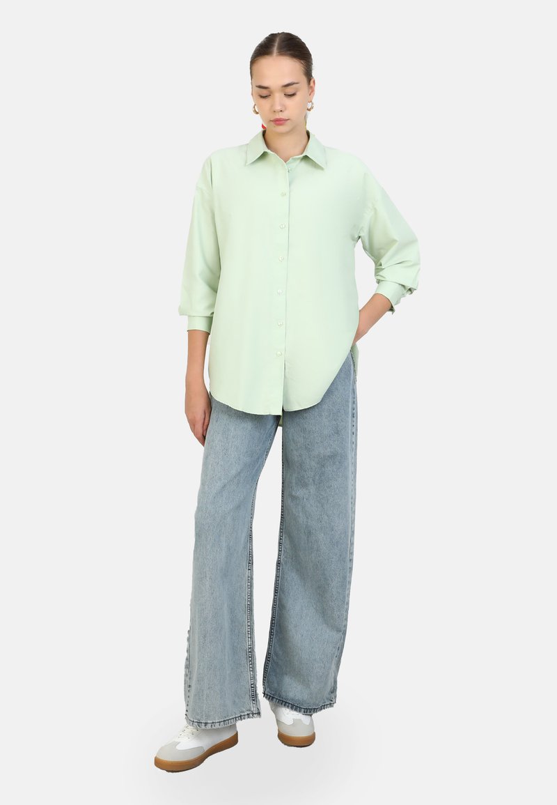 Camisa de botonadura en un verde claro con cuello, mangas largas y corte relajado, combinada con jeans de pierna ancha en azul claro y zapatillas blancas.