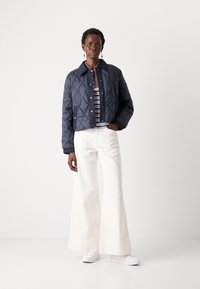 Gequiltede marineblauwe jas met een kraag, drukknoopsluiting en geribde manchetten, gecombineerd met witte wijde broek en witte sneakers.