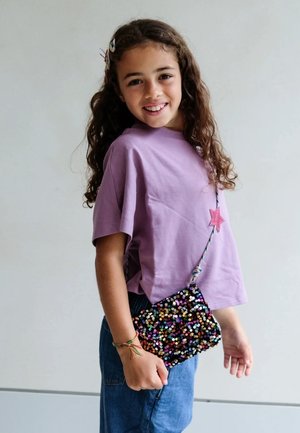 Camicetta corta viola con dettaglio a stella rosa abbinata a jeans blu. Tiene in mano una piccola borsa a tracolla nera con paillettes e dettagli colorati.