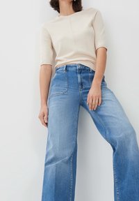 Beige lyhythihainen paita ja vaaleansiniset leveälahkeet farkut. Paidassa on sileä pinta; farkuissa on haalistunut pesu ja muotoillut taskut.