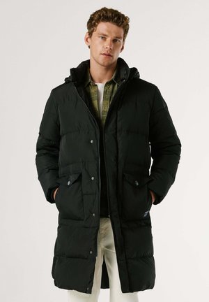 Młody mężczyzna w długim czarnym pikowanym płaszczu, na zielonej koszuli i jasnych spodniach, stoi z rękami w kieszeniach płaszcza.