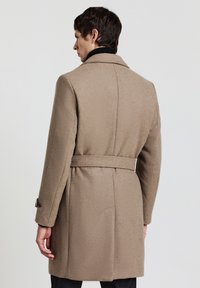 Cappotto in lana beige con cintura, ampio colletto e polsini abbottonati, caratterizzato da un tessuto testurizzato e una vestibilità su misura nella parte posteriore.