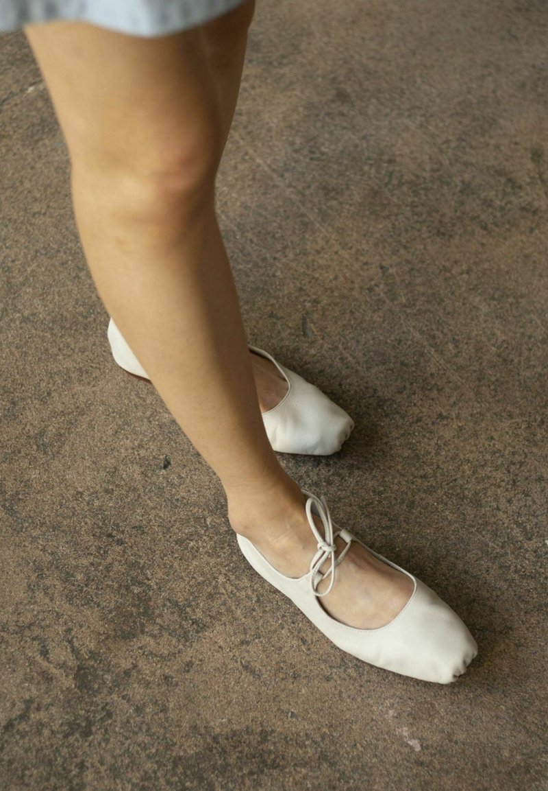 Ballerine beige chiaro realizzate in morbido cuoio, con punta arrotondata e un sottile laccio decorativo davanti, appoggiate su una superficie testurizzata.
